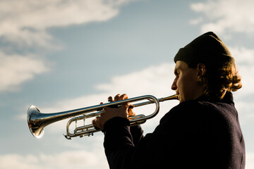A man with a hat playing a trumpet isolated on blue cloudy sky. Male Jazz musician play music on...