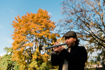 A man with a hat playing a trumpet on autumn yellow tree background. Male Jazz musician play music...