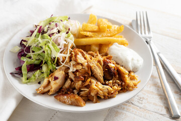 Doner kebab chicken meat with french fries, salad and tzatziki dip on a white plate, cutlery and napkin on a rustic wooden table, selected focus