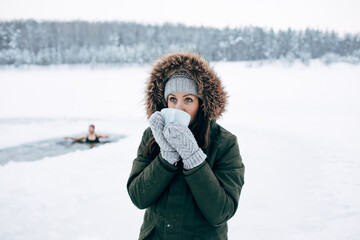 Beautiful young female smiling and drinking tea in nature Winter swimming. Woman in frozen lake ice...