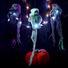 Halloween decoration three skeletons in a multicolored cape decorated with a garland of glowing pumpkins and spiders. There is a decorative pumpkin at the bottom