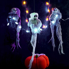 Halloween decoration three skeletons in a multicolored cape decorated with a garland of glowing pumpkins and spiders. There is a decorative pumpkin at the bottom