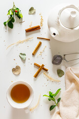 Composition with tea pot, cup  of tea, cinnamon, tea strainer, linen fabric, green plant leaves on white background. Flat lay, top view. 
