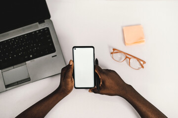 African American man holding smart phone with mockup white blank display, empty screen for app ads sitting on a couch at home. Mobile applications technology concept, over-the-shoulder close up view.