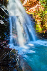 Detail of waterfall over cliffs with red rocks and moss into blue bucket of water