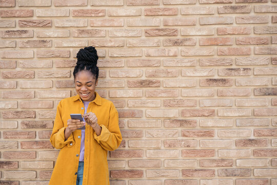 Young Beautiful African American Woman Using Mobile Phone Shopping Online Standing Near Wall, Copy Space. Smiling Hipster Female Wearing Stylish Yellow Shirt Holding Smartphone Communication Online