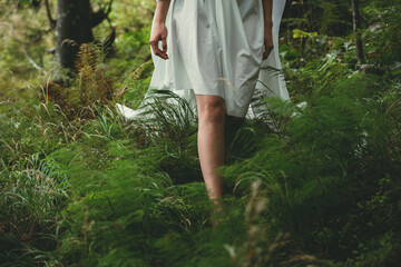 legs of a young girl who walks barefoot in a white dress on green grass in the forest on a sunny day, front view close-up