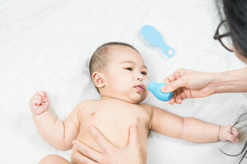 Mom holding her body to a small boy with her hands sucks the snot from the baby's nose lying on the bed through the aspirator. Cleaning the nose on the bed.Baby treatment, colds, immunity, cleansing.