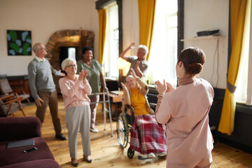 Diverse group of smiling senior people dancing while enjoying activities in retirement home with young woman in foreground, copy space