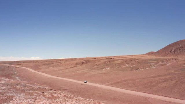 Aerial dolly shot of white car disappearing along a red dirt road in the the Atacama Desert in Chile, South America near the salt lakes of Laguna Baltinache, red and orange mountains in the distance