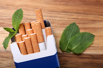 Pack of menthol cigarettes and mint leaves on wooden table, flat lay