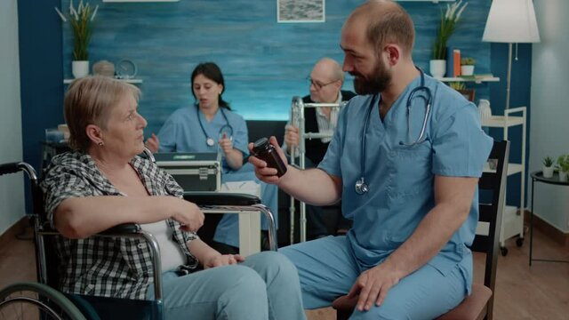Man nurse giving bottle of pills to old patient with disability for healthcare treatment in nursing home. Senior woman in wheelchair receiving flask with medicine for medical recovery