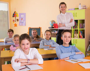 Smiling children sitting at lesson in classroom, teacher standing