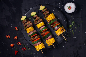 Beef skewers with red onion, green zucchini, red paprika and sweet cob with barbecue salt and thyme on a black background.