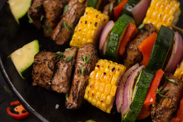 Beef skewers with red onion, green zucchini, red paprika and sweet cob with barbecue salt and thyme on a black background.