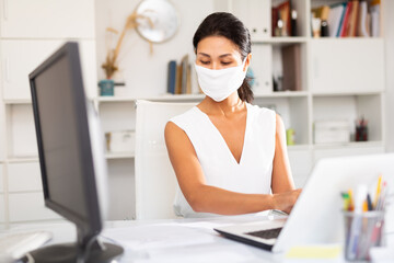 Young businesswoman in medical mask and white dress working with laptop and papers in office....