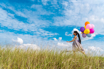 Obraz premium Cheerful cute girl holding balloons running on green meadow white cloud and blue sky with happiness. Hands holding vibrant air balloons play on birthday party happy times summer on sunlight outdoor