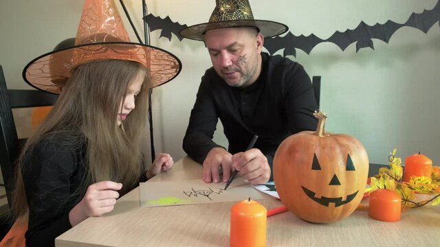 Child with father draw pictures on the theme of halloween in decorated room at home. Little girl and her dad smiling, laughing and having fun together, happy family preparing to celebrate the holiday.