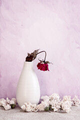 Single dried rose in a vase against a painted purple and white background; Simple design of red flower in a curved vase surrounded by dried soft flowers