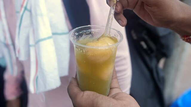 A person eating Kanji Vada made with flavored water - Served in plastic glass. People queueing up and waiting outside a famous Indian Chaat shop in Old Delhi - street food