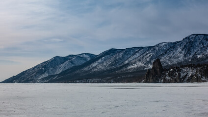 The frozen lake is covered with snow. Coastal wooded mountain range against the blue sky. Winter Baikal