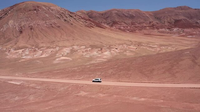 Aerial dolly shot of white car driving along a red dirt road in the the Atacama Desert in Chile, South America near the salt lakes of Laguna Baltinache, red and orange mountains in the distance