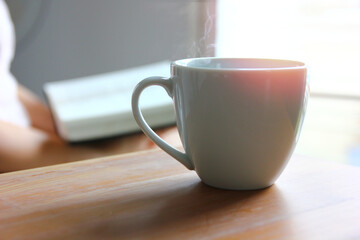 hot coffee in white ceramic cup on wood table with woman reading book background in morning light