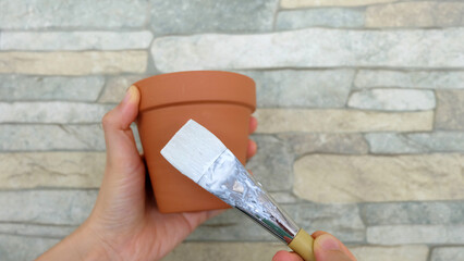 Hand holding a new terracotta pot, with a paint brush with white paint placed over the pot.