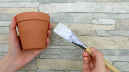 Hand holding a new terracotta pot, with another hand holding a white paint brush near the pot.