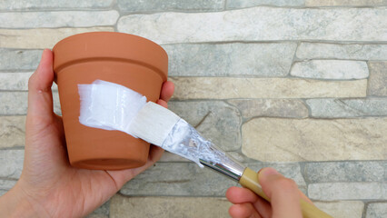 Hand holding a new terracotta pot, with another hand holding a white paint brush, making a stroke on the pot.