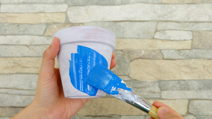 Hand holding a terracotta pot with white base paint, with another hand holding a blue paint brush, coloring the pot with blue paint.