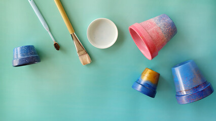 Flat lay of colorful painted terracotta pots, paintbrush, toothbrush and a paint tray. With copy space on the bottom left corner.