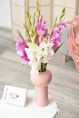 Vase with beautiful gladiolus flowers and picture frame on table in room