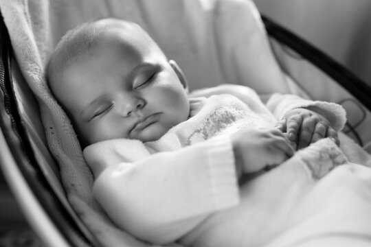Cute baby laying in bouncer chair and sleep. Child relaxing in a swing. Adorable newborn baby in bodysuit. Family morning at home. Selective focus. Black and white photo.