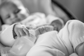 Cute baby laying in bouncer chair and sleep. Child relaxing in a swing with milk bottle. Adorable newborn baby in bodysuit. Family morning at home. Selective focus. Black and white photo. 