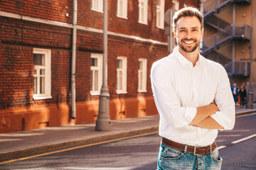 Fototapeta premium Portrait of handsome smiling stylish hipster lambersexual model.Modern man dressed in white shirt. Fashion male posing on the street background. Outdoors at sunset