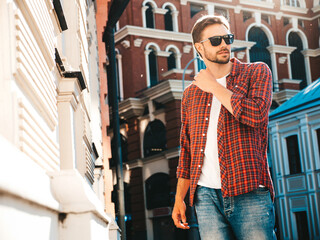 Obraz premium Handsome confident stylish hipster lambersexual model.Modern man dressed in red checkered shirt. Fashion male posing near skyscraper on the street background in sunglasses. Outdoors at sunset