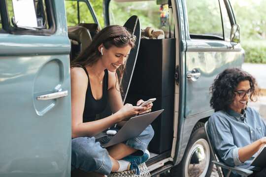 Smiling woman using smart phone while male friend with book sitting by outside camping van