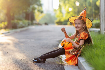 Lifestyle portrait of Happy Little caucasian Girl with blonde hair seven years old in black orange costume of which celebrating Halloween alone outdoor during Coronavirus covid-19 pandemic quarantine.