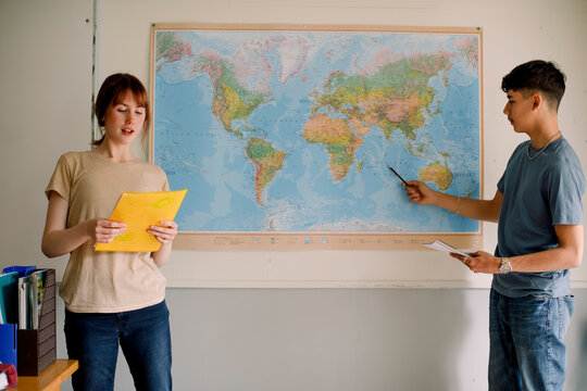Teenage girl reading while boy pointing on world map in classroom