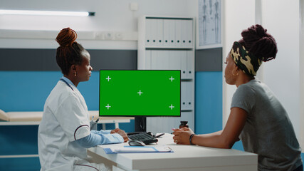 Medical specialist looking at horizontal green screen on computer monitor. Doctor working with chroma key for isolated mockup template and background on display, talking to patient.