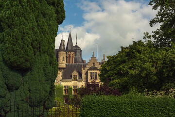 Impressive summer garden with trees, hedges, varied plants and an iron gate with a historic castle under a blue cloudy sky.