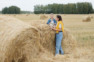 Naklejka na ściany i meble Beautiful young mother and cute son toddler are having fun on a haystack in the field 