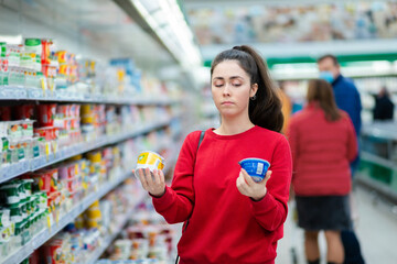 A young caucasian woman stands in a supermarket and thoughtfully chooses between two yogurts....
