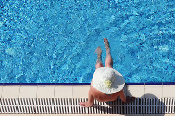 Woman in sun hat and bikini sitting on the edge of swimming pool and dangling her legs in water, top view. Vacation on beach resort, tanning and leisure concept