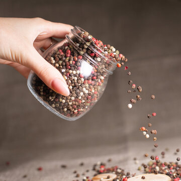 Crop woman pouring spicy oriental peppercorn mix from glass jar on table on gray background 