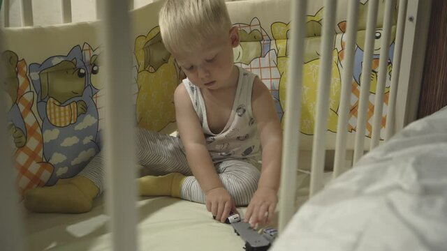 A fair-haired preschool boy playing with toy cars while sitting in a crib. Portrait of a kid doing leisure activity. Close-up. Home bedroom interior.