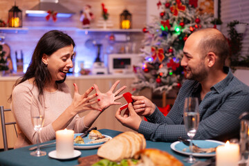 Happy man surpsing girlfriend with diamond luxury engagement ring enjoying christmas holiday together sitting at dinning table in xmas decorated kitchen. Married couple enjoying winter season