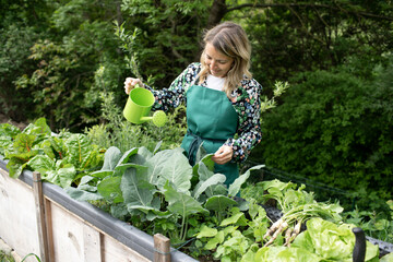 young blonde woman watering vegetables in garden with small green watering can and is happy