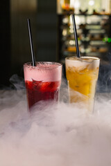 Red and orange alcohol cocktails in glasses on bar counter with smoke in nightclub 